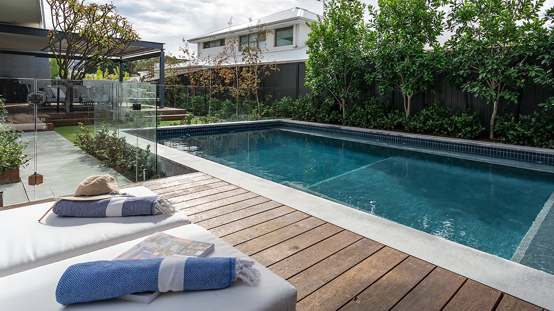 a contemporary landscape with a timber deck beside a tiled concrete pool, the pool is lined with layered trees and hedges.